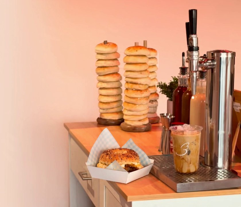 A Baglory counter set-up: stacked bagels on wooden pegs, a boxed bagel sandwich, an iced coffee and a nitro tap.