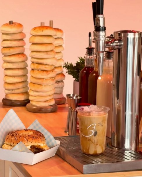 A stainless nitro tap tower beside an iced coffee with the Baglory heart and stacks of bagels on wooden pegs.